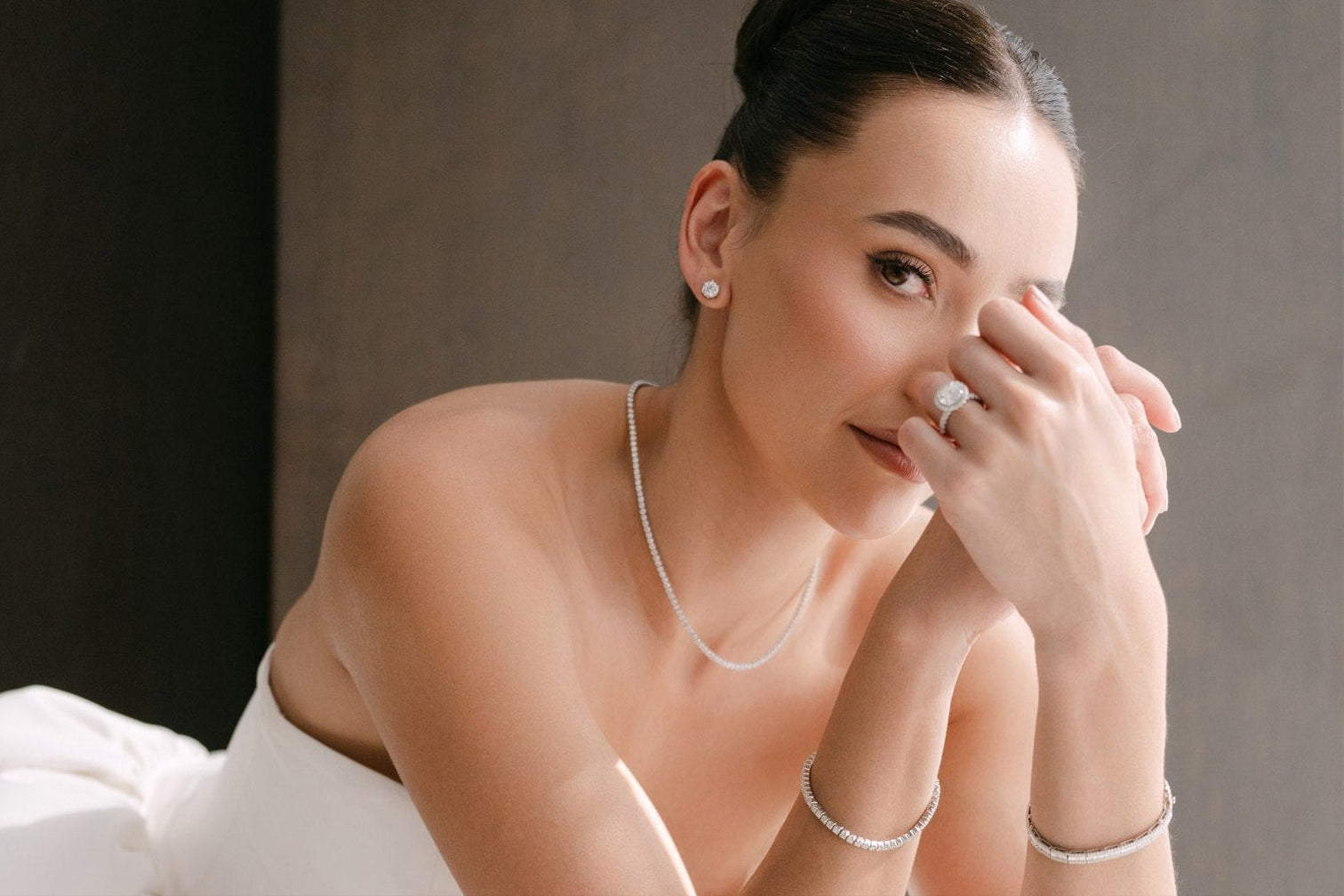 Woman in a white dress with jewelry, sitting on a white surface against a neutral background